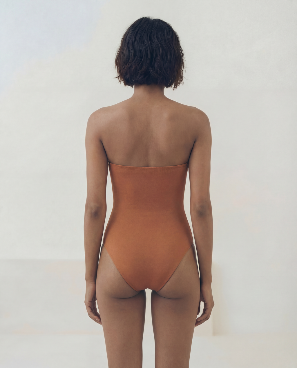 A woman viewed from behind wearing a strapless bandeau-style one-piece swimsuit in a warm terracotta or rust orange color, set against a plain off-white studio background.