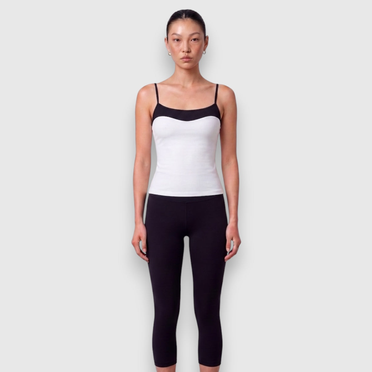 A model wearing a white camisole with black trim and black leggings, standing against a light gray background.