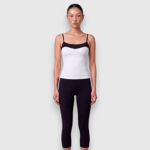 A model wearing a white camisole with black trim and black leggings, standing against a light gray background.