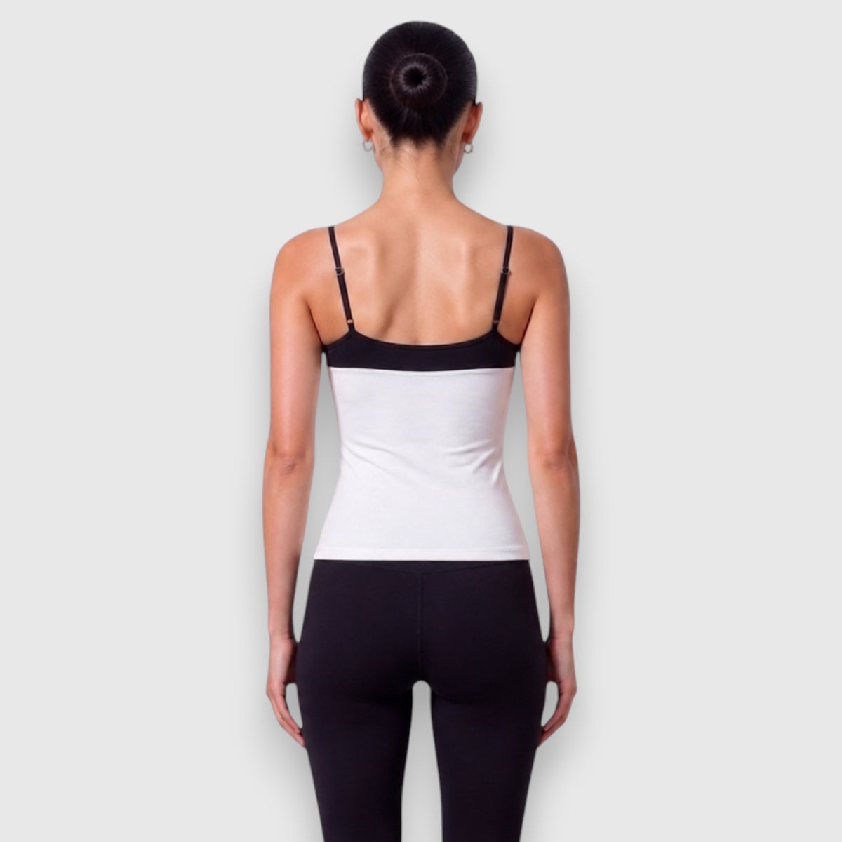 A woman viewed from the back wearing a white camisole-style top with black shoulder straps and a black band across the upper back, paired with black leggings, against a light gray studio background.